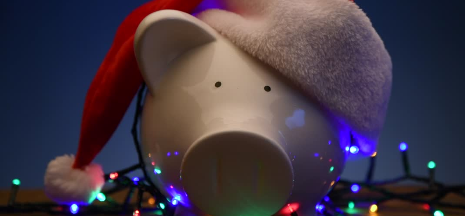 piggy bank wearing a red and white santa hat. multicolor Christmas lights on the surface. 